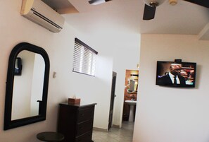 A hotel room with a large mirror, a dresser, an air conditioning unit, a television, a window with blinds, and a bathroom visible in the background. - Altamont West Hotel (Montego Bay)