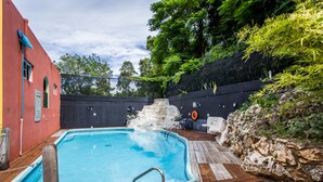 Une piscine dotée d’une terrasse en bois, entourée d’un mur de pierre et de verdure.
