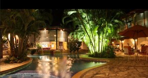 Un espace piscine aménagé pour la nuit, avec un élément décoratif aquatique, des palmiers et des sièges d’extérieur.