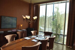 A conference room with a wooden table, patterned chairs, a chandelier, a painting, and a view of trees through large windows.