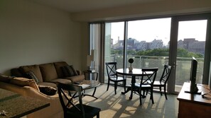 A living room with a sofa, coffee table, and chairs, a TV on a stand, and a balcony with a city view. - Charles River Executive Suites (Boston)