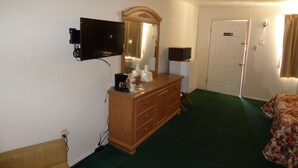 A hotel room with a wooden dresser, a flat-screen TV mounted on the wall, a bedside lamp, a door, and a green carpet. - Carrizozo Inn (Carrizozo)