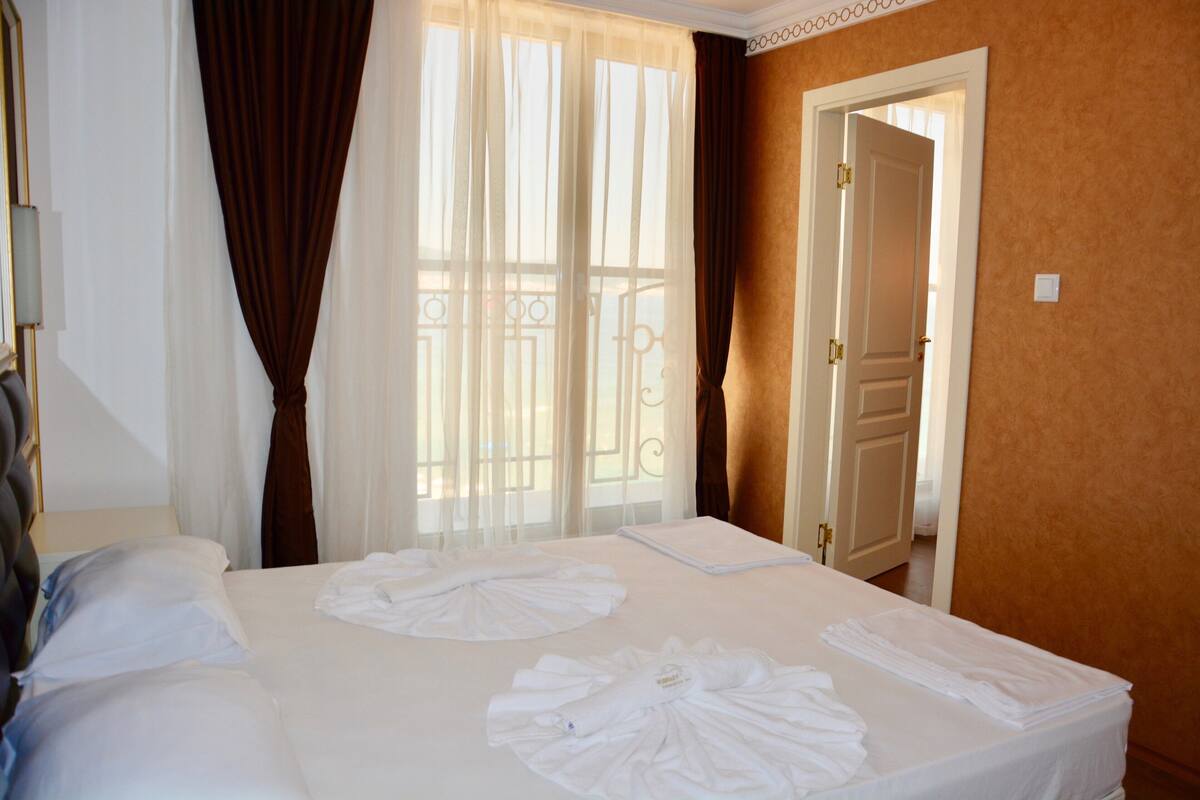 A hotel room with a large bed, a bedside table, a window with curtains, and a door.