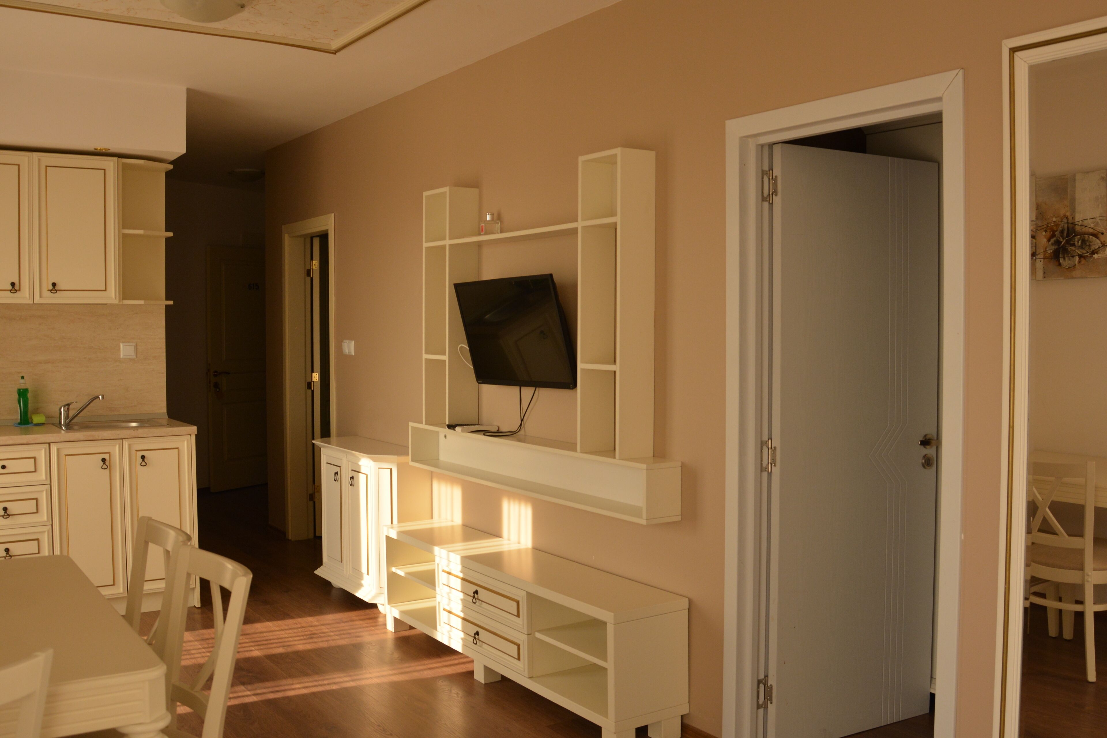 Habitación con televisión montada en un mueble blanco, una mesa de comedor con sillas y una puerta que da a otra estancia.