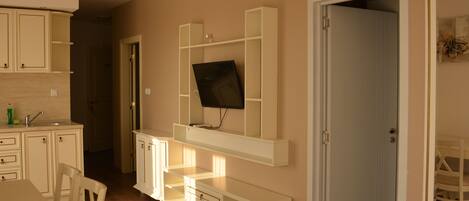 Habitación con televisión montada en un mueble blanco, una mesa de comedor con sillas y una puerta que da a otra estancia.