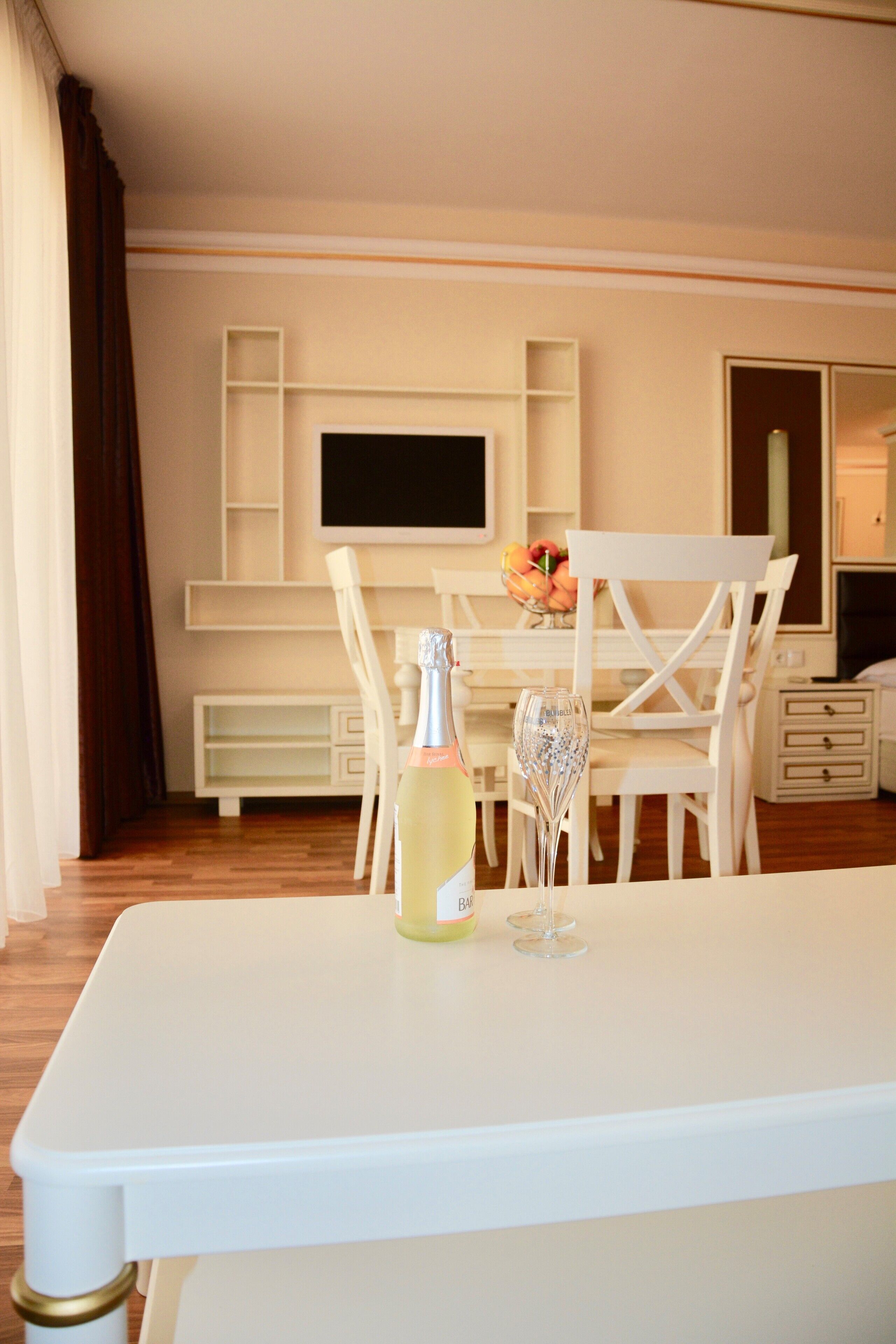 A hotel room with a white table, a bottle of champagne, and a glass on the table. There is a TV mounted on the wall and a white chair in front of the TV.