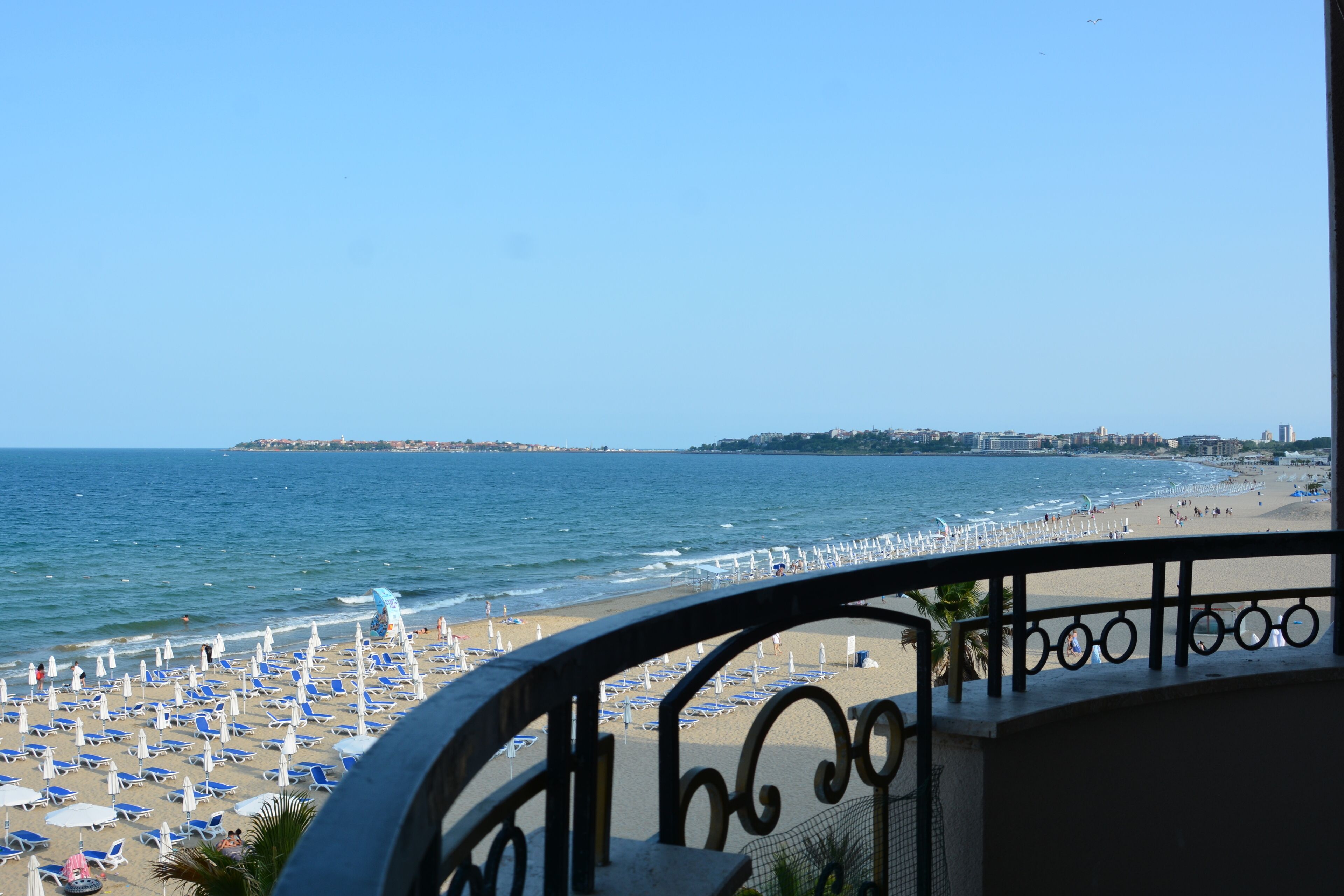 Une plage bordée de rangées de transats et de parasols, un ciel dégagé et une vue sur la mer et les bâtiments éloignés.