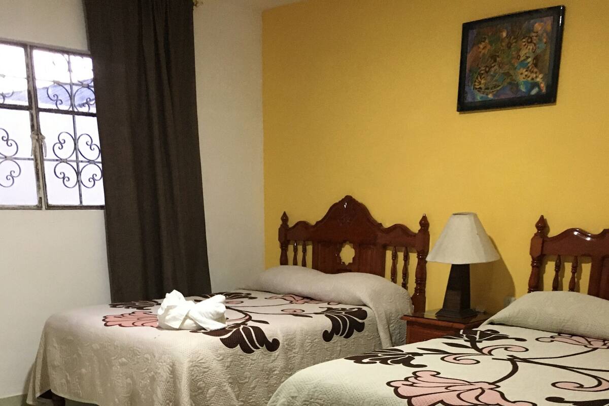 A room with two beds, a window with curtains, a nightstand with a lamp, and a painting on the wall.