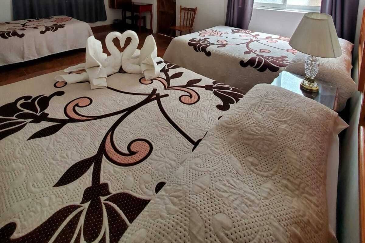 A hotel room with two beds, a small table with a lamp, a window with curtains, and a ceiling light.