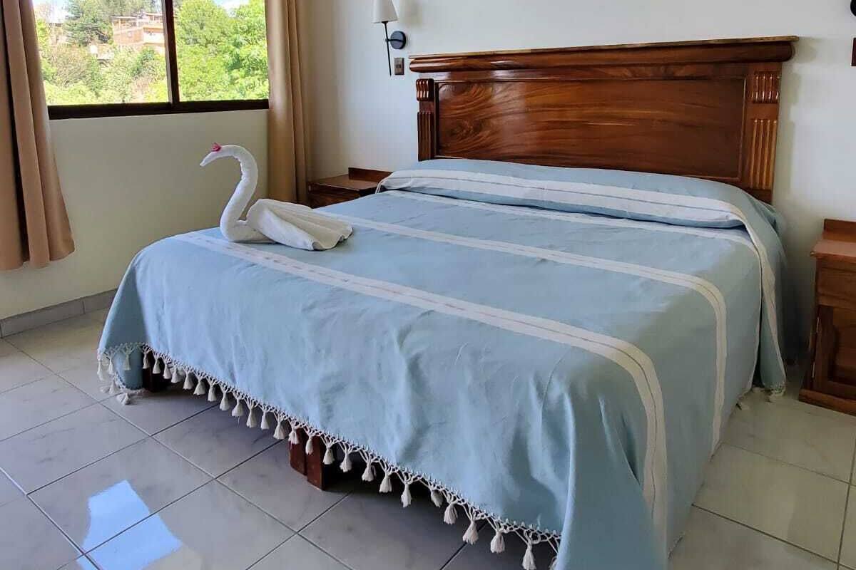 A bedroom with a large bed, a wooden headboard, a window with curtains, and a view of greenery outside.
