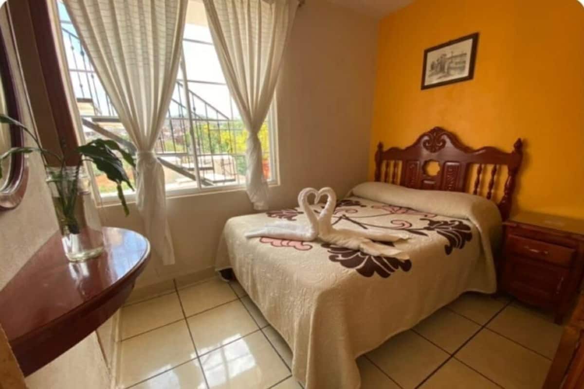 A bedroom with a bed, a wooden headboard, a bedside table, a window with curtains, and a wall-mounted picture.