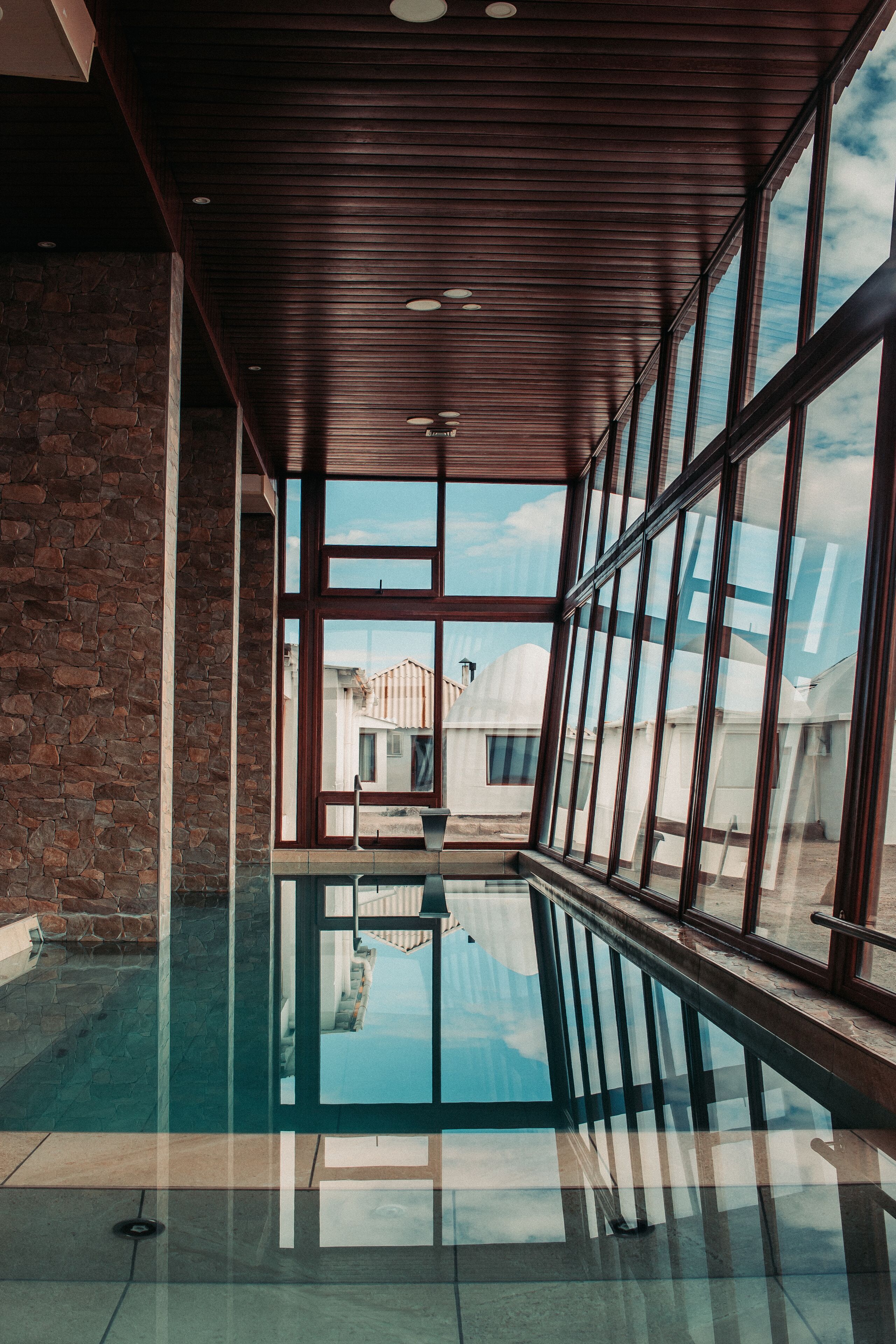 An indoor swimming pool with a glass floor, wooden ceiling, and large windows.