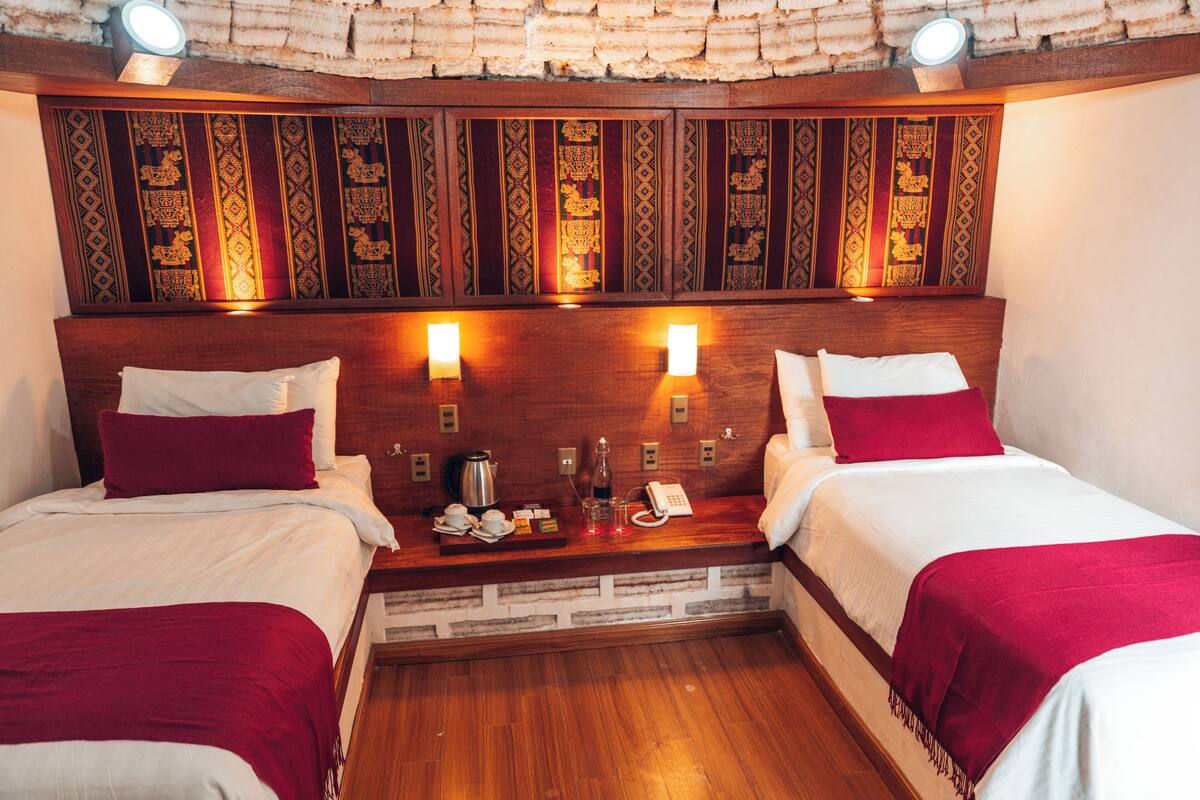A hotel room with two beds, a wooden headboard with intricate patterns, bedside lamps, a nightstand with a phone, a kettle, and a tray with cups and a teapot.