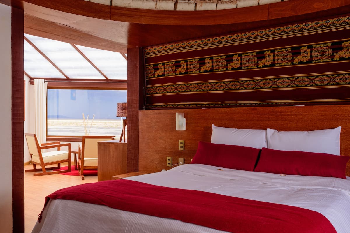A hotel room with a large bed, a wooden headboard with intricate designs, a desk with a chair, and a view of the beach through a large window.