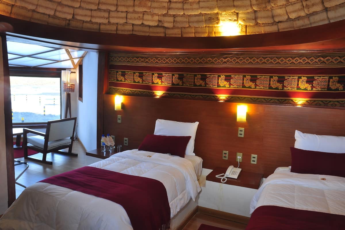 A hotel room with two beds, a wooden headboard with decorative patterns, bedside lamps, and a view of the outdoors.