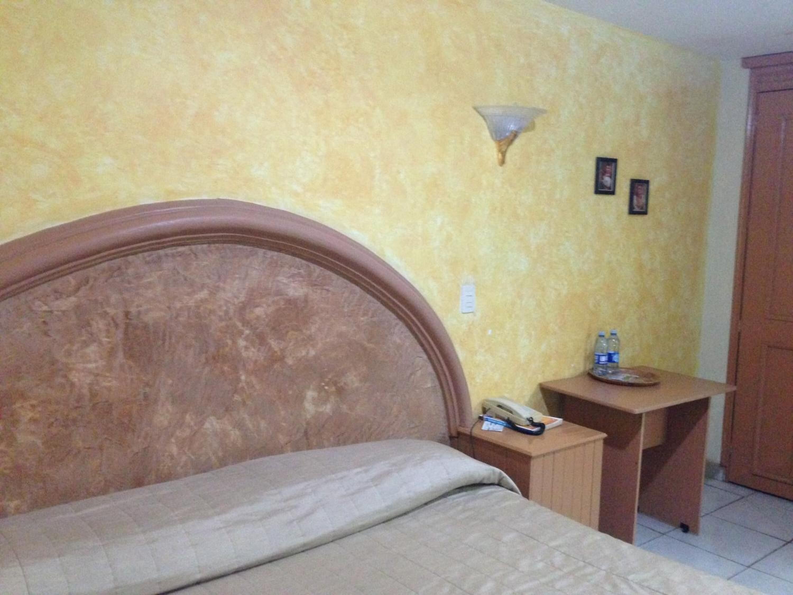 A bed with a wooden headboard, a bedside table with a lamp, and a small table with a phone and water bottles.