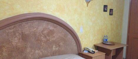 A bed with a wooden headboard, a bedside table with a lamp, and a small table with a phone and water bottles.