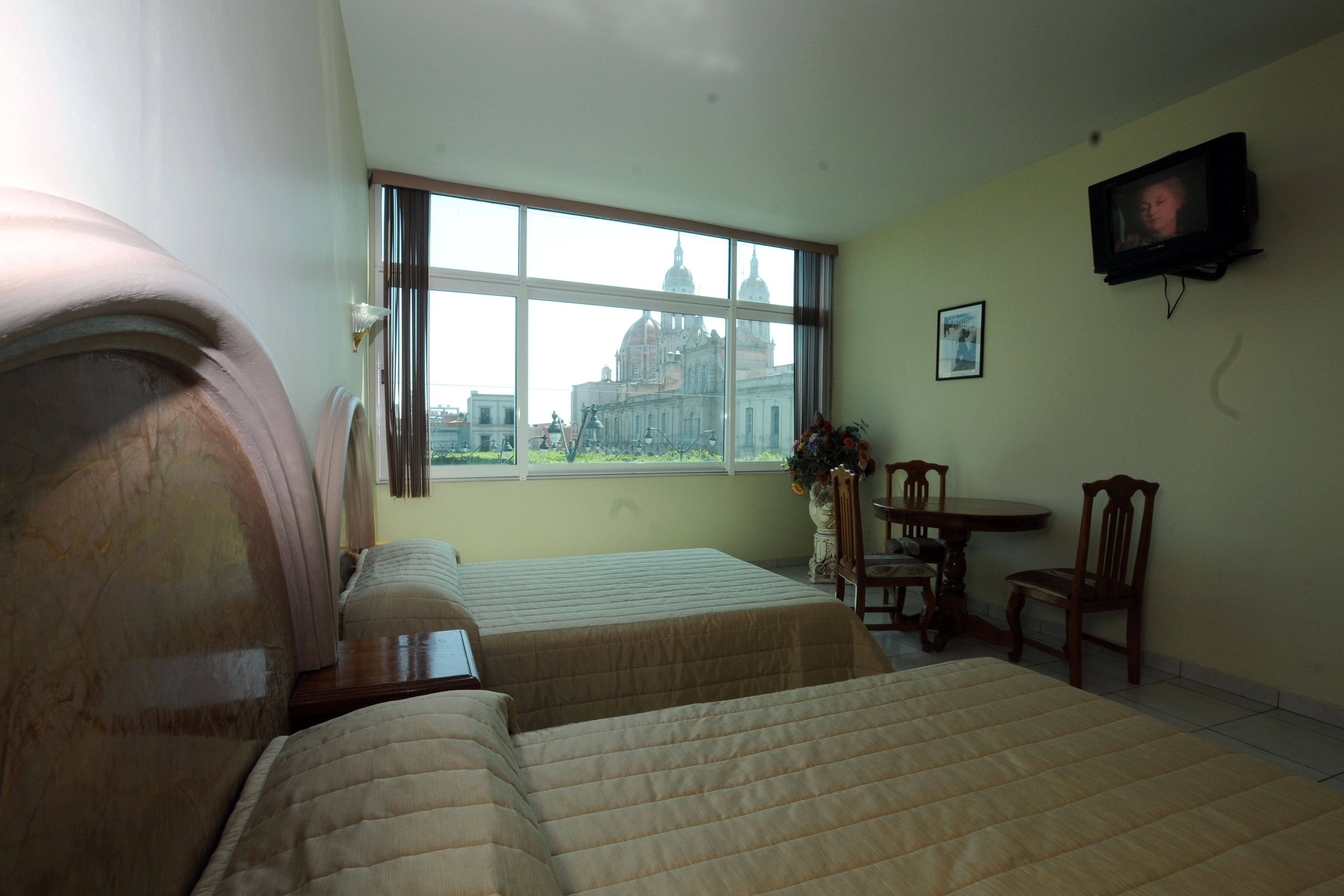 Camera d'albergo con due letti, televisione, una finestra vista su un edificio e un piccolo tavolo con sedie.