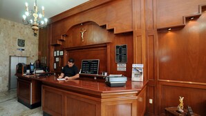 Un comptoir d’accueil en bois, agrémenté d’un lustre, d’une plaque portant un crucifix et d’un panneau indiquant les tarifs des chambres.