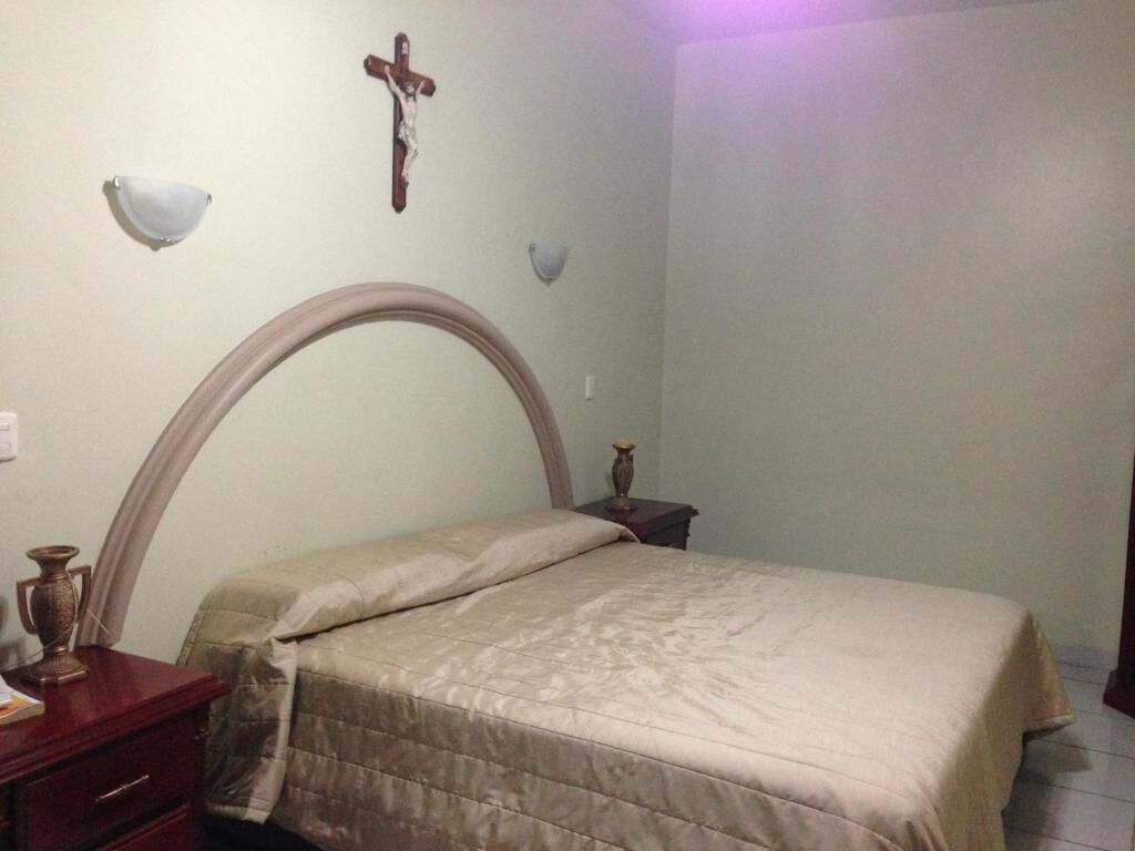 Un dormitorio con una cama, una mesita de noche, una cruz en la pared y un estante fijado a la pared.
