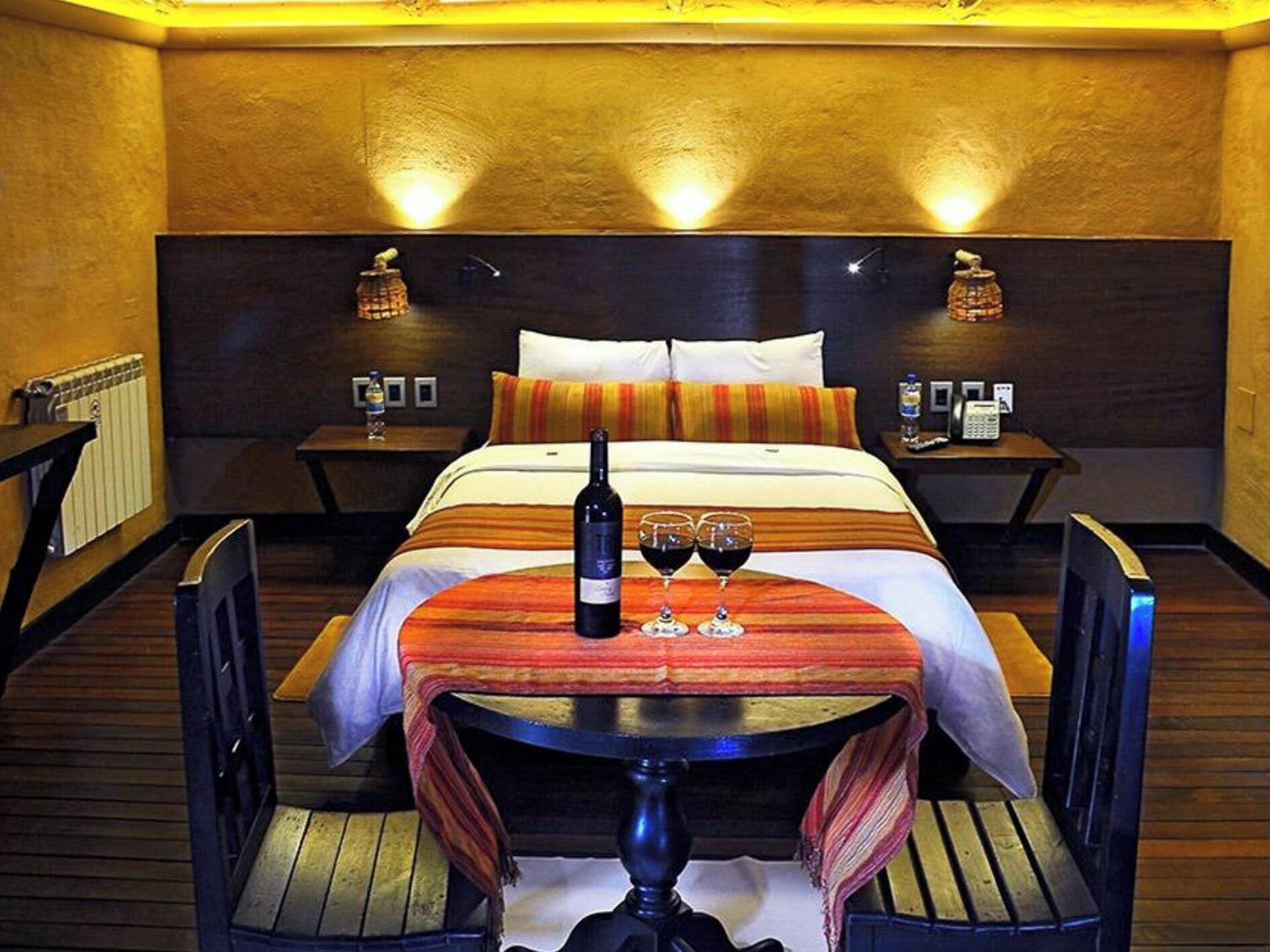 Une chambre d’hôtel avec un lit, des tables de chevet, une table avec des verres à vin, une bouteille et une chaise.