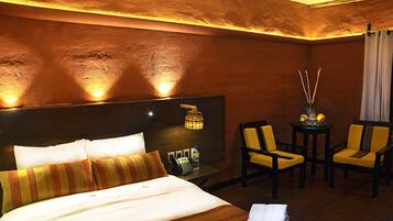 A hotel room with a bed, bedside table, lamp, two chairs, and a small table with a vase and fruit.