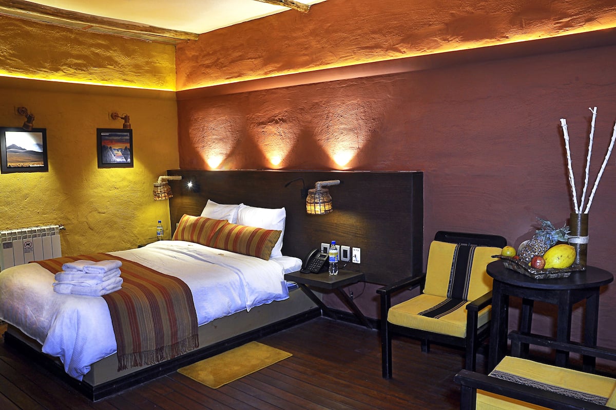 Een hotelkamer met een bed, een nachtkastje met telefoon en waterfles, een stoel en een tafeltje met fruit en een decoratief object.