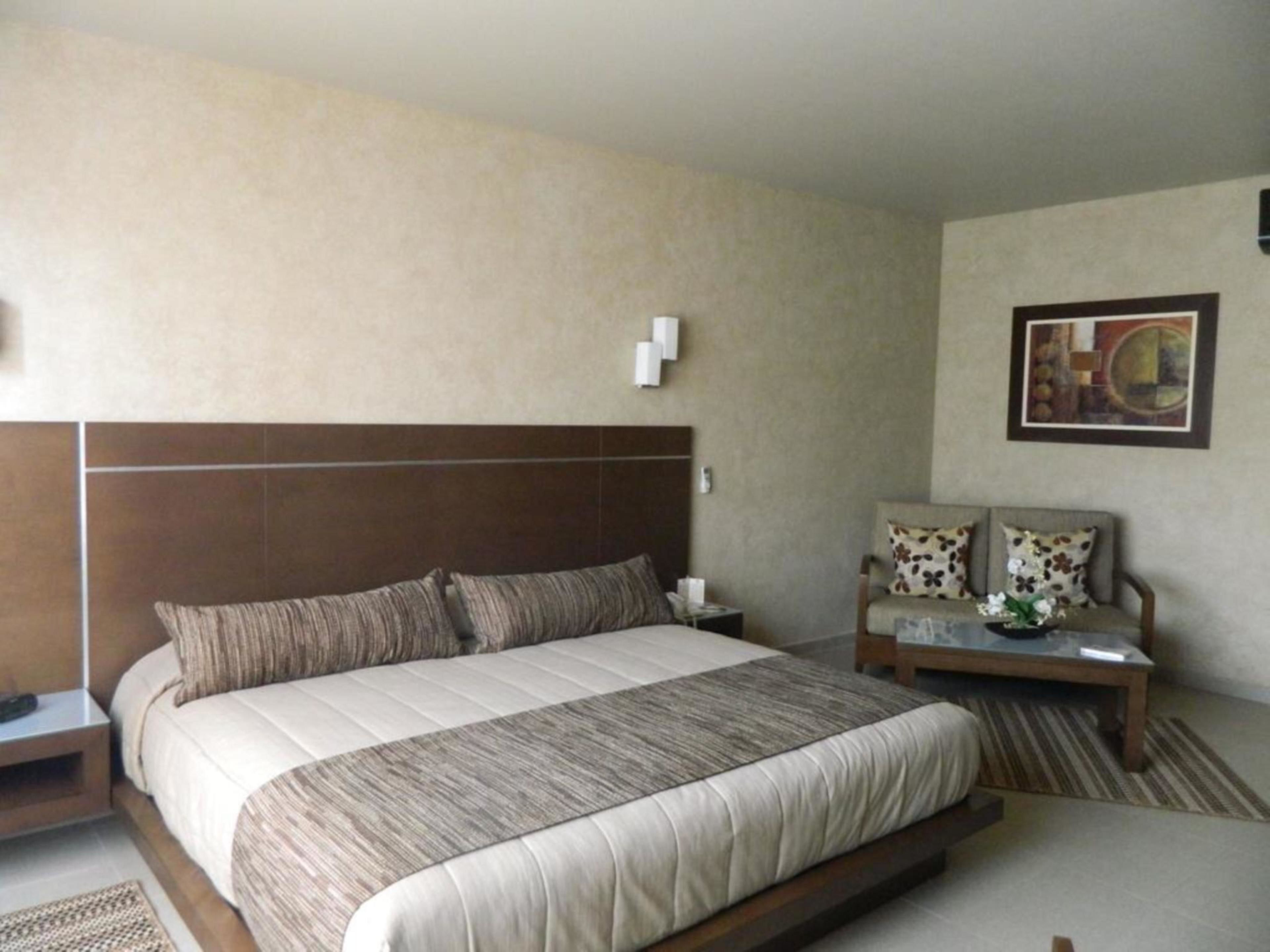 Habitación de hotel con una cama grande, una mesita de noche con lámpara, una silla y una pequeña mesa con un florero.