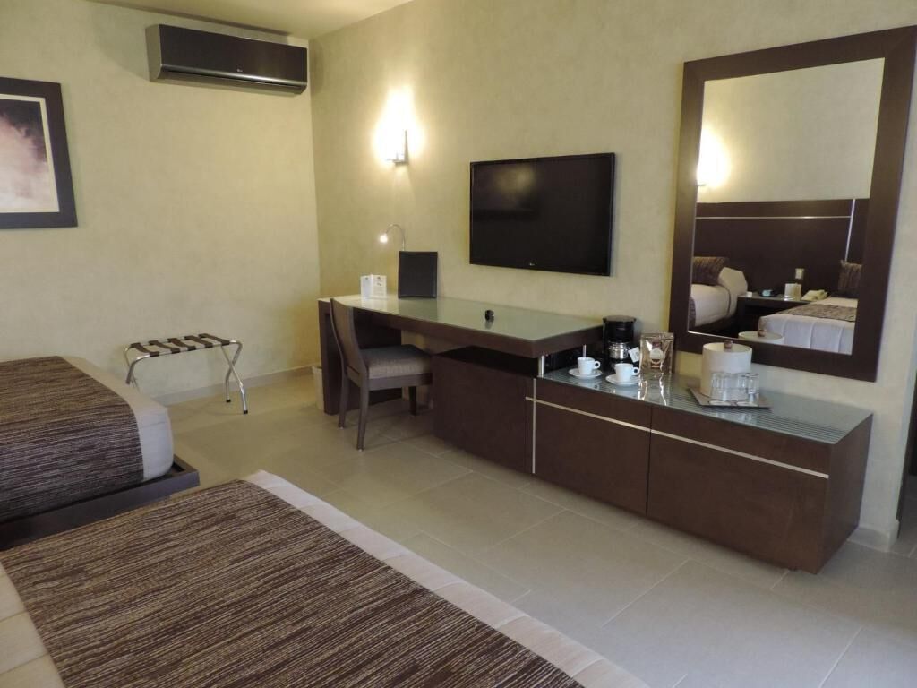 Habitación de hotel con cama, escritorio, silla, televisión, espejo y cafetera.