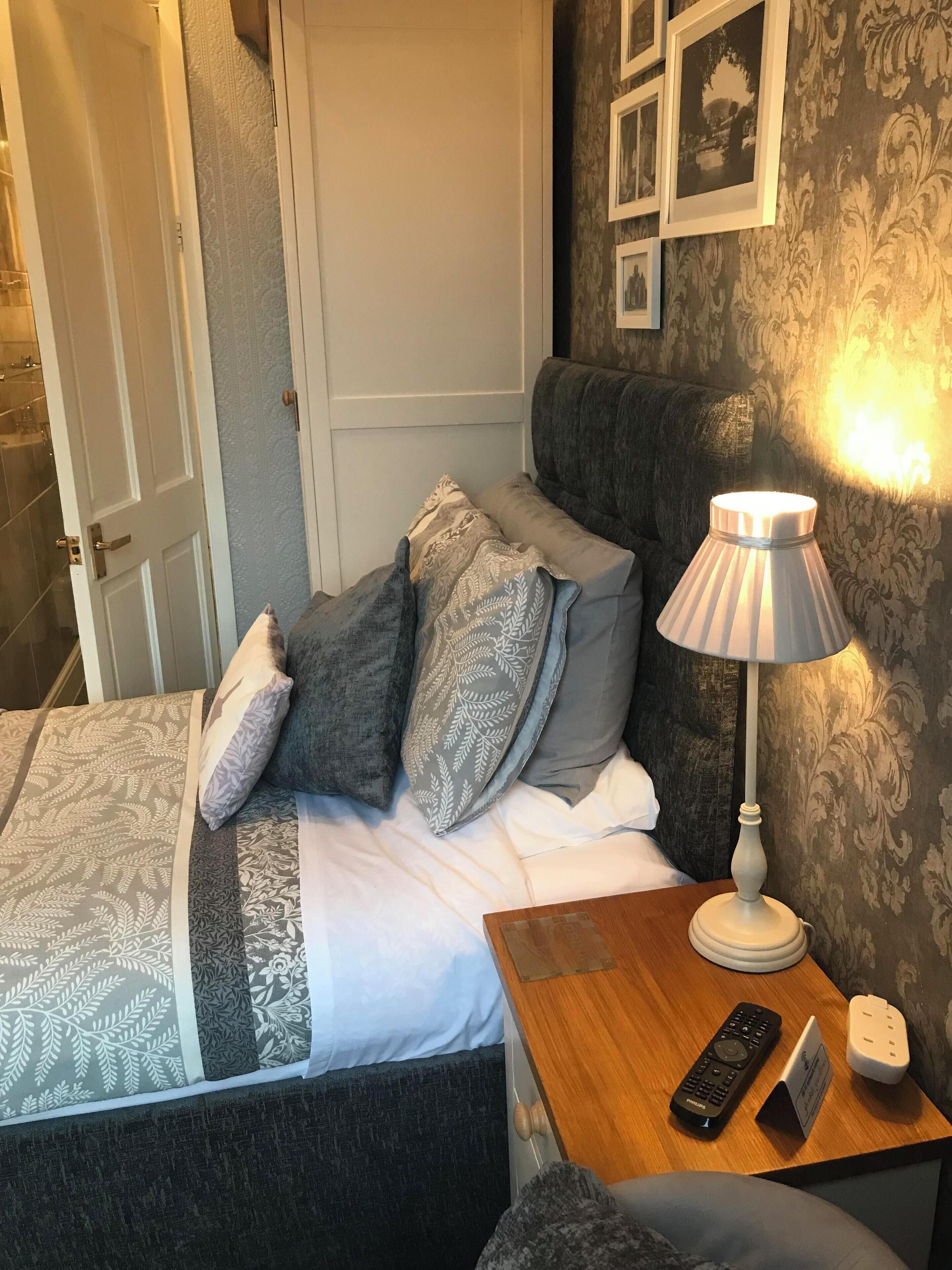 Een net opgemaakt bed met behang, een bedlampje, een afstandsbediening en een oplaadstation op een nachtkastje van hout.
