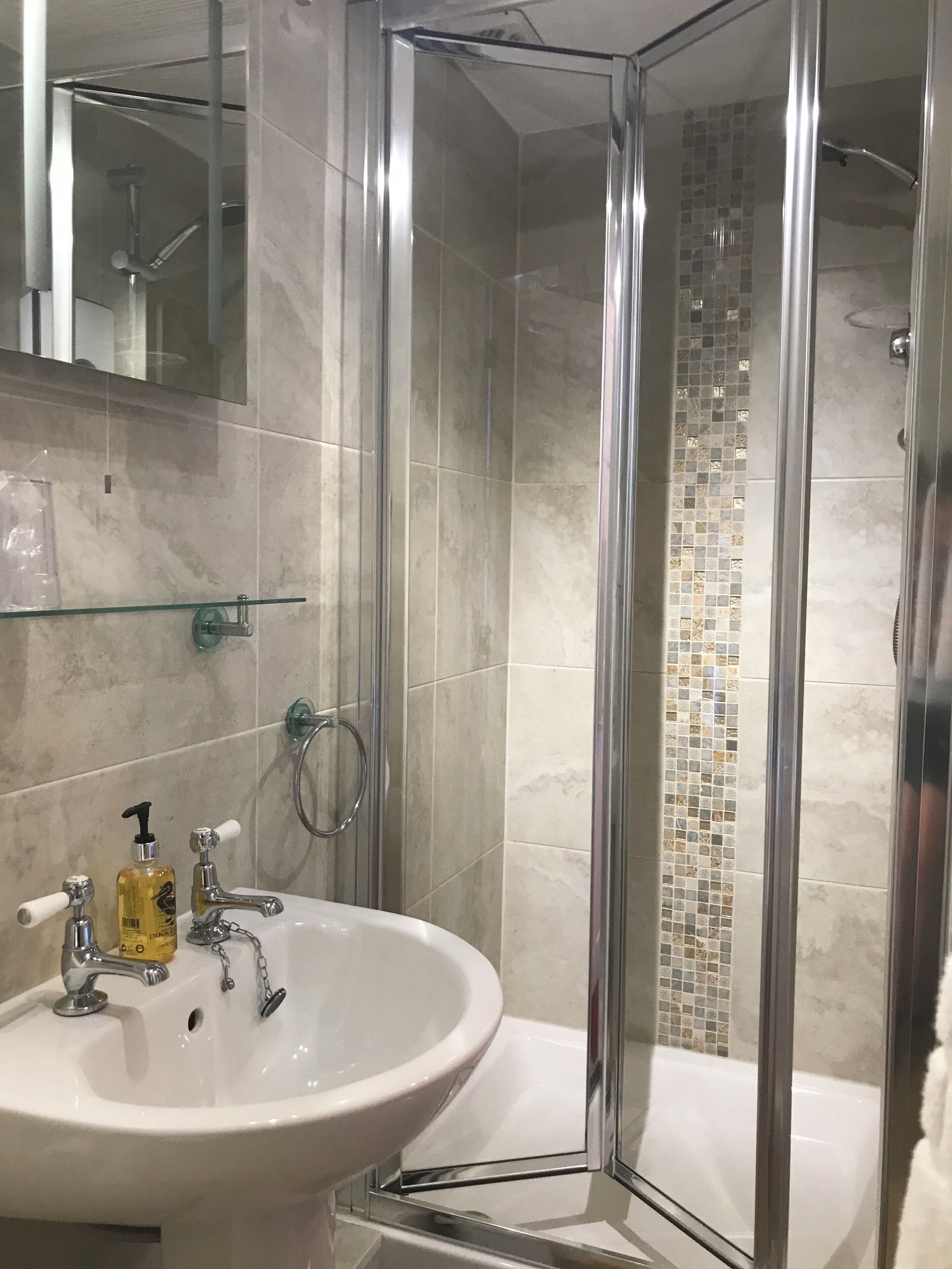 Ein modernes Badezimmer mit einer Duschkabine, einem weißen Waschbecken mit Chromarmaturen und einer gefliesten Duschwand.