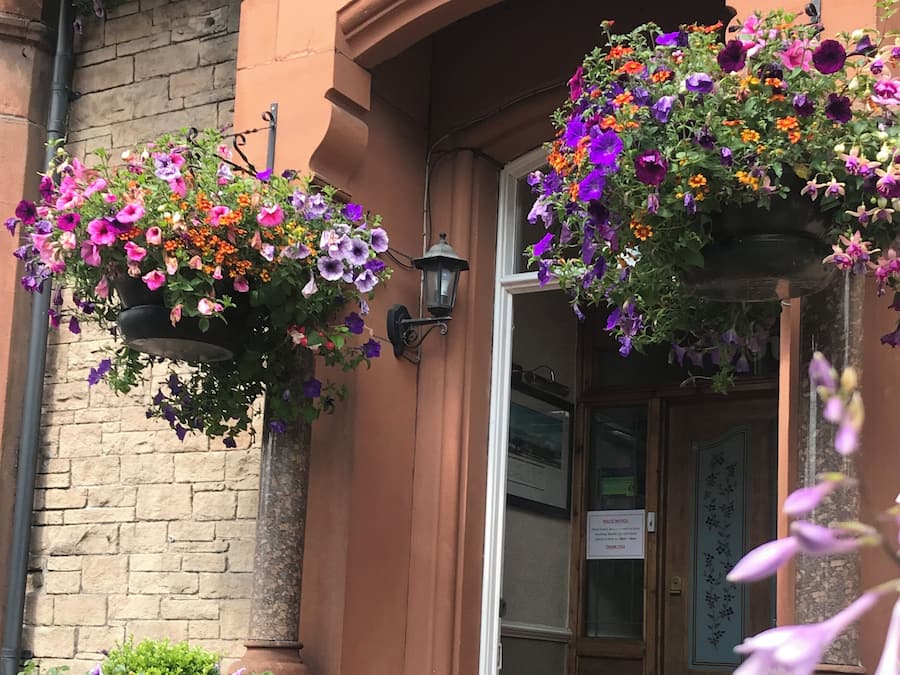 Entrada de un edificio con cestas de flores colgantes y una puerta con un letrero.