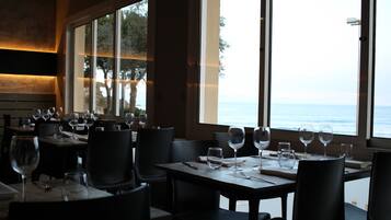 Un restaurante con mesas dispuestas para comer, grandes ventanales con vistas al mar.