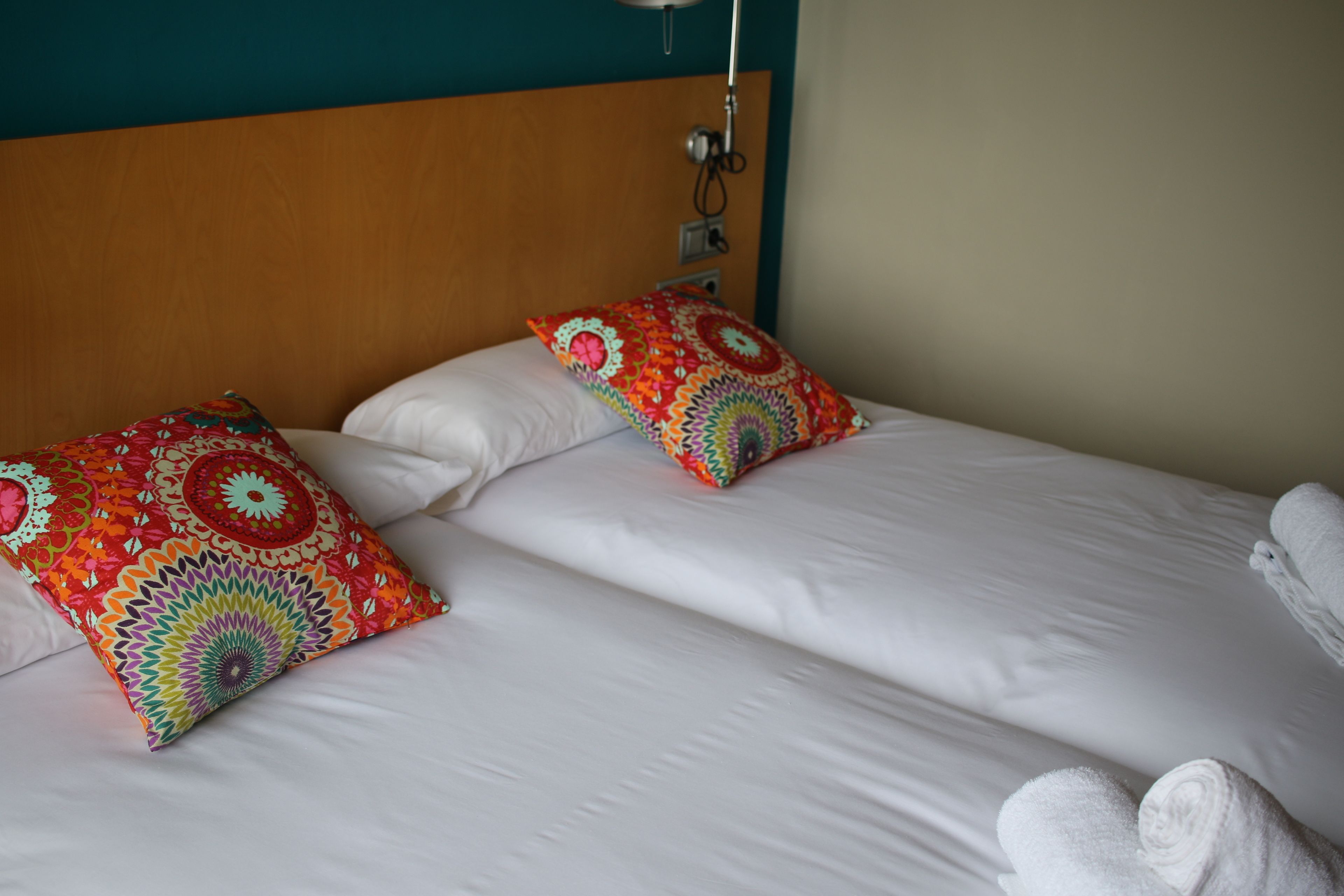 A hotel room with a wooden headboard, two beds with white linens, colorful patterned pillows, and a rolled white towel on the right side.