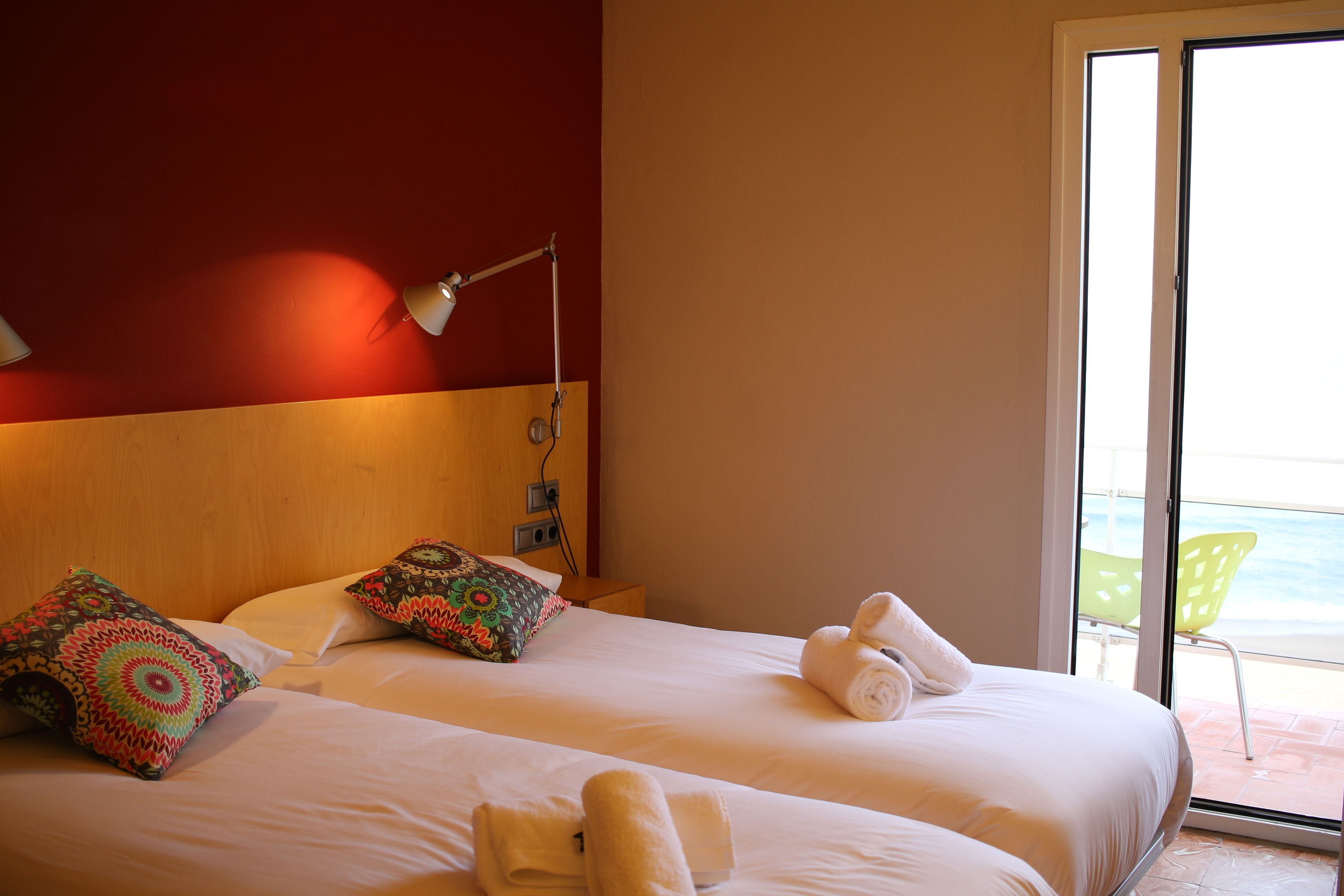 A hotel room with two beds, a wooden headboard, a nightstand, a lamp, and a view of the sea through a sliding glass door.