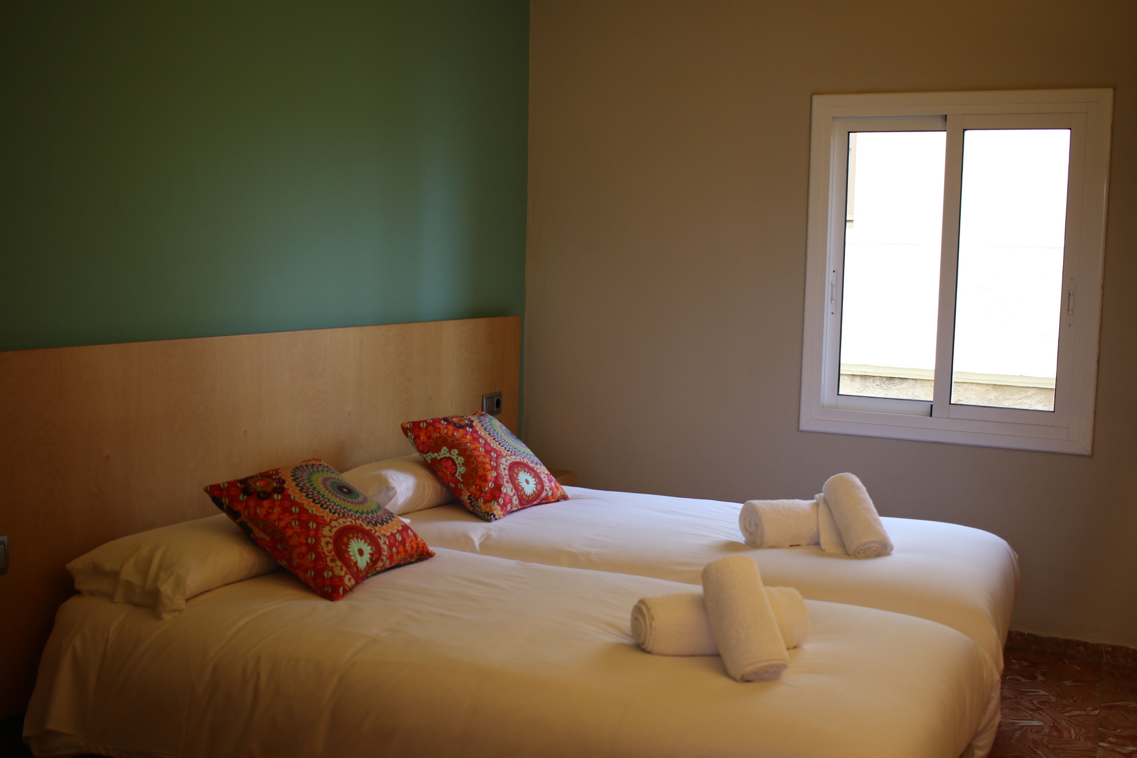 Un lit double avec une literie blanche et des coussins colorés, une fenêtre avec des stores et une tête de lit en bois.