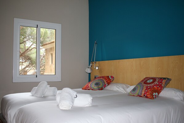 A neatly made bed with white linens and two decorative pillows, a window with a view of trees, and a bedside lamp.