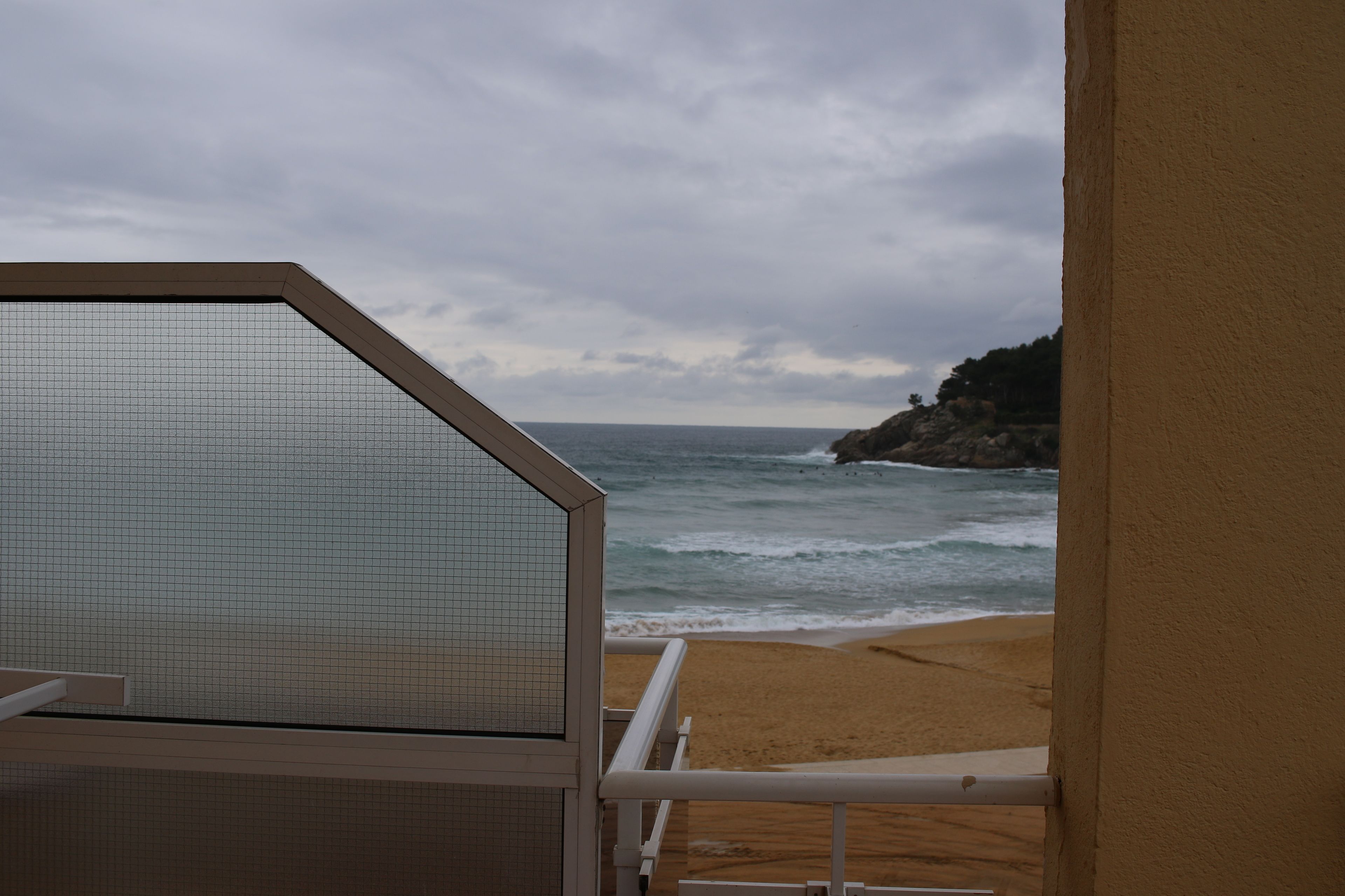 A beach view from a balcony with a railing, a sandy beach, and rocky cliffs in the background.