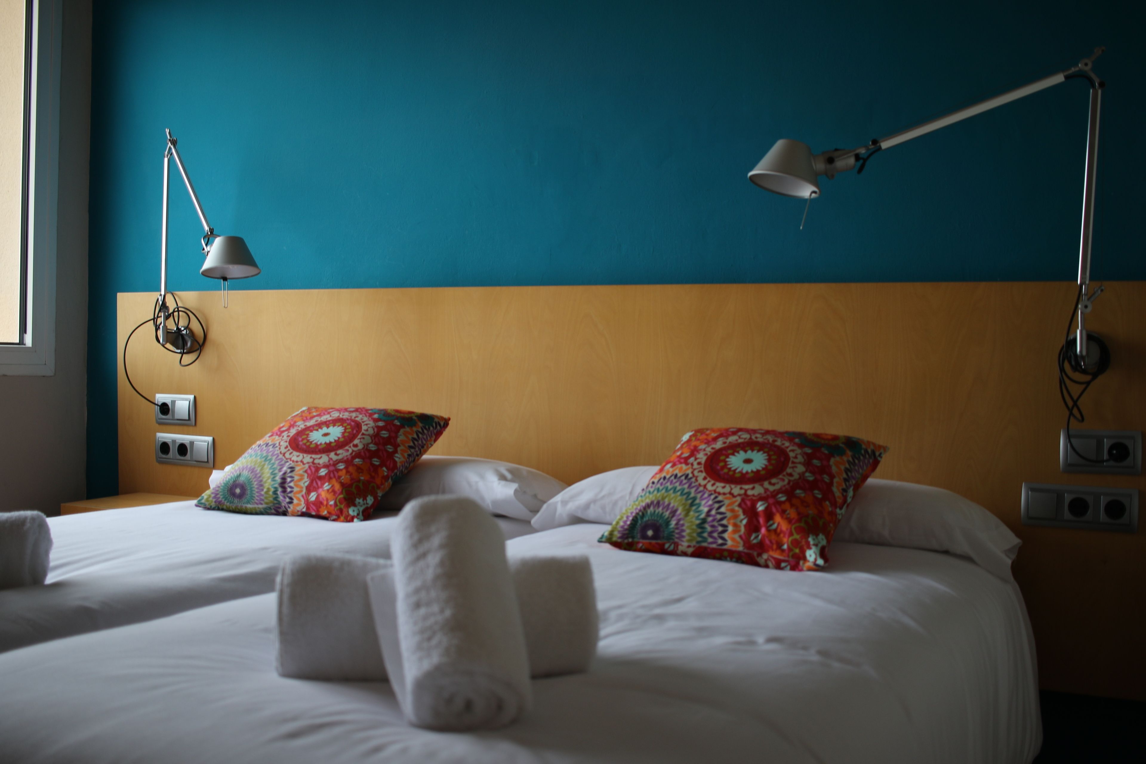 Una habitación de hotel con una cama, dos almohadas coloridas, una manta blanca y dos lámparas de noche.