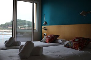 A hotel room with a large window offering a view of the sea and hills. There are two beds with white linens and colorful pillows. - Hostal LA Fosca (Palamós)