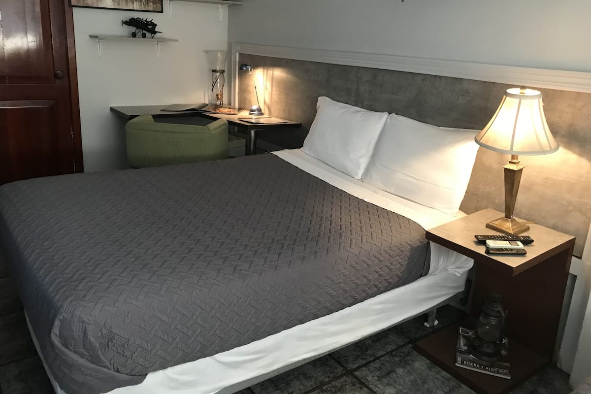 A neatly made bed with a grey quilt, a bedside lamp, and a small table with a lamp and remote controls.