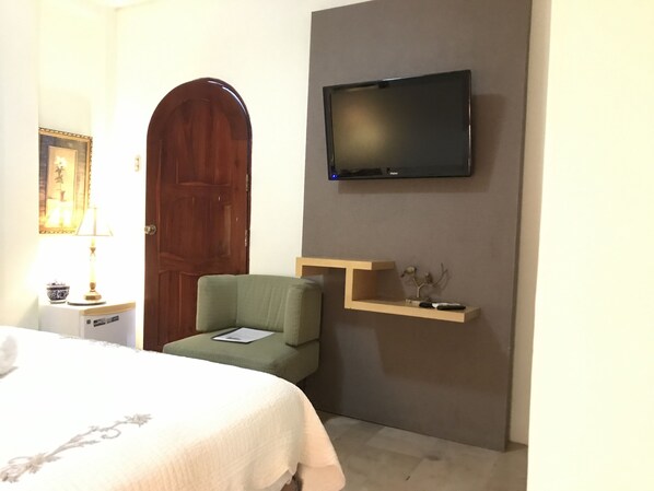 Una habitación de hotel con una cama, un sillón verde, un televisor de pantalla plana en la pared, una puerta de madera y un pequeño estante con objetos decorativos.