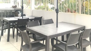 An outdoor patio area with tables and chairs, a large umbrella, and a view of greenery and buildings in the background. - K7 SUITES (Samborondon)