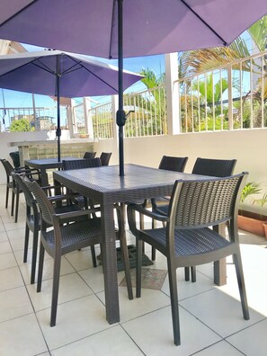 En udendørs patio med et bord og stole, en lilla parasol samt udsigt til palmer og bygninger.