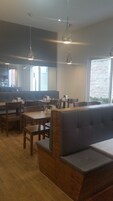 Un restaurante con mesas y sillas de madera, un banco acolchado gris y lámparas colgantes, además de amplias ventanas con vistas al exterior.