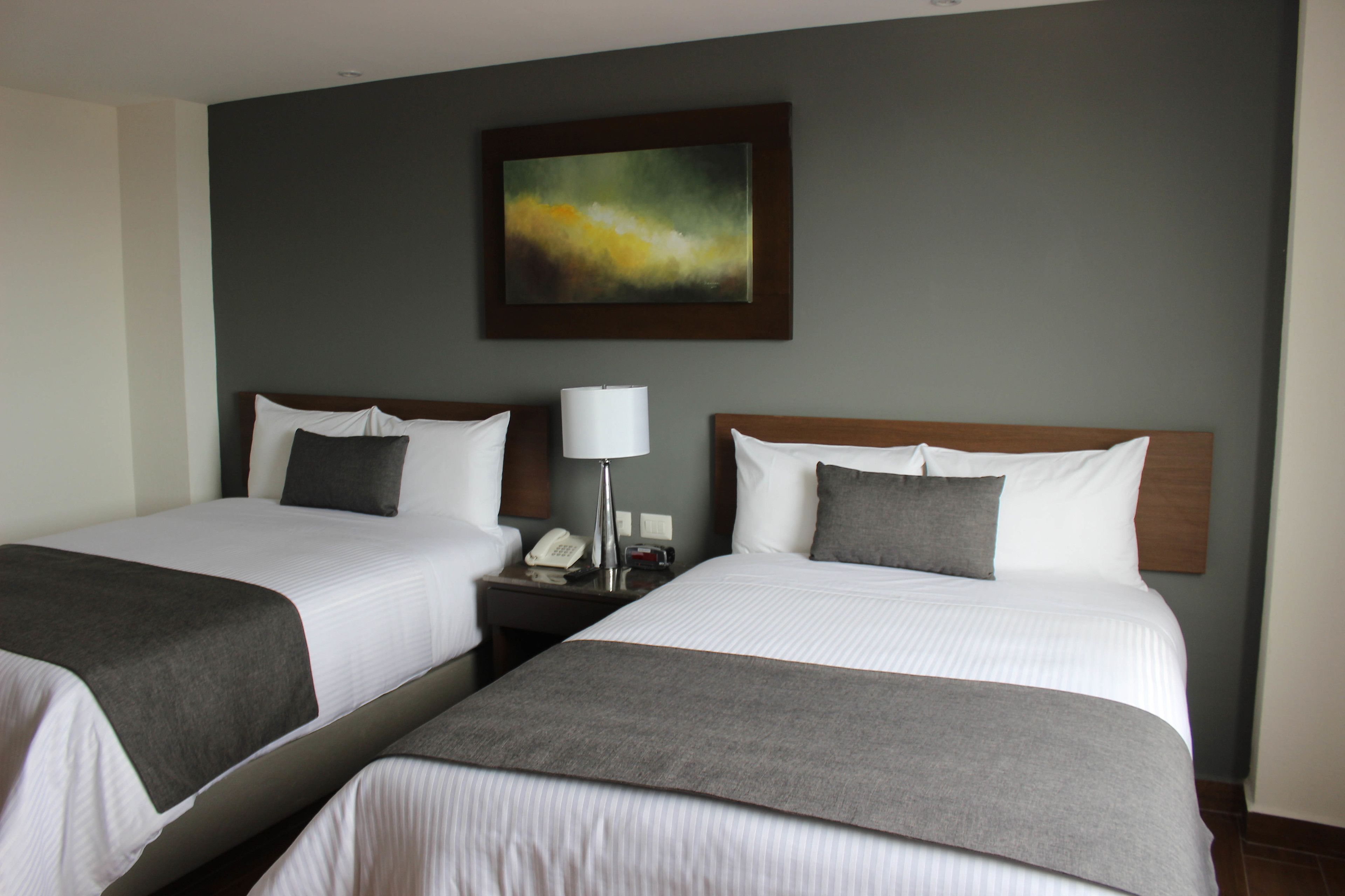 Une chambre d’hôtel avec deux lits, chacun étant agrémenté de draps blancs et d’un plaid gris. Une table de chevet avec une lampe et un téléphone se trouve entre les lits. Un tableau est accroché au mur, au-dessus des lits.