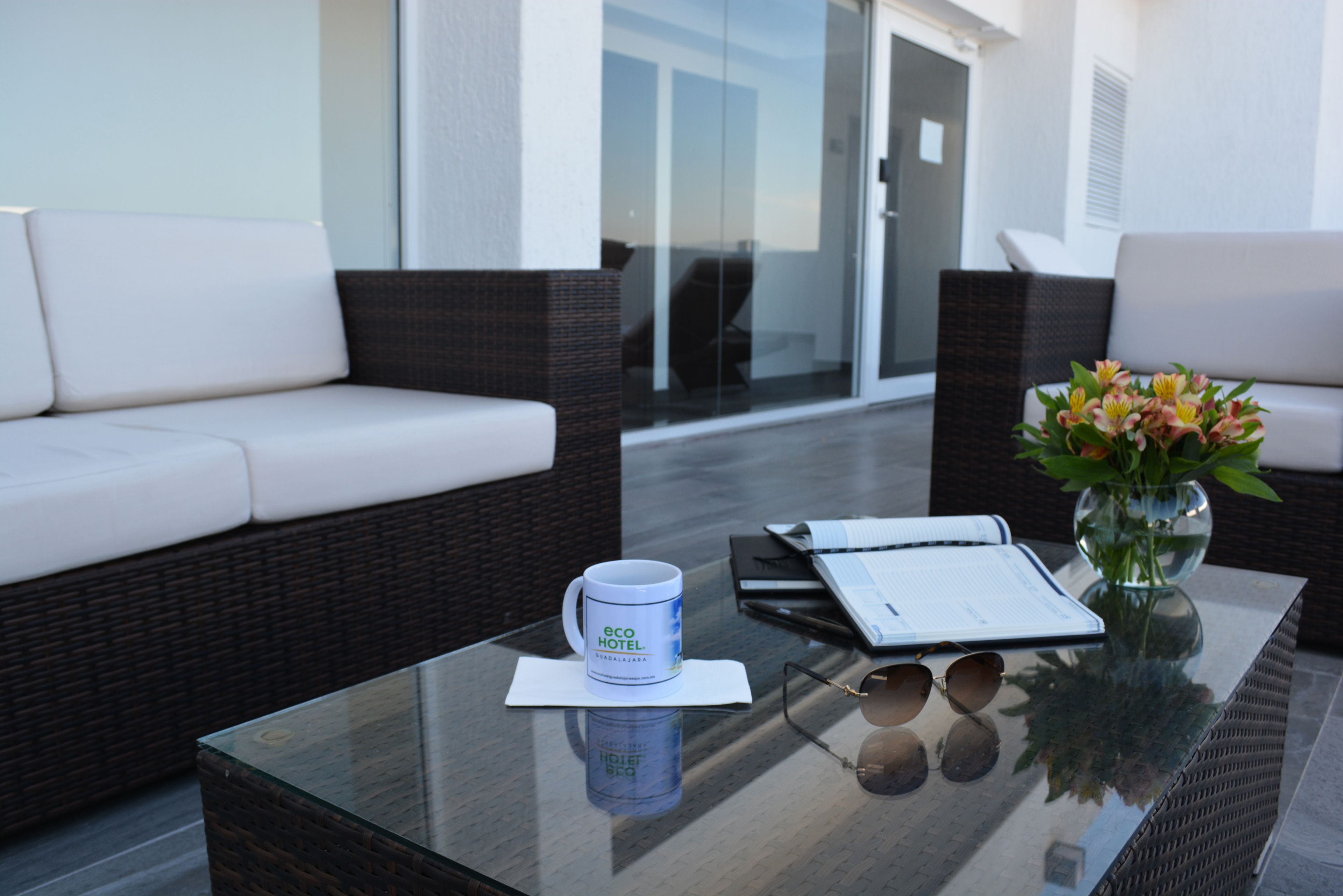 Una mesa con cubierta de vidrio, una taza blanca, unos lentes de sol y un cuaderno.