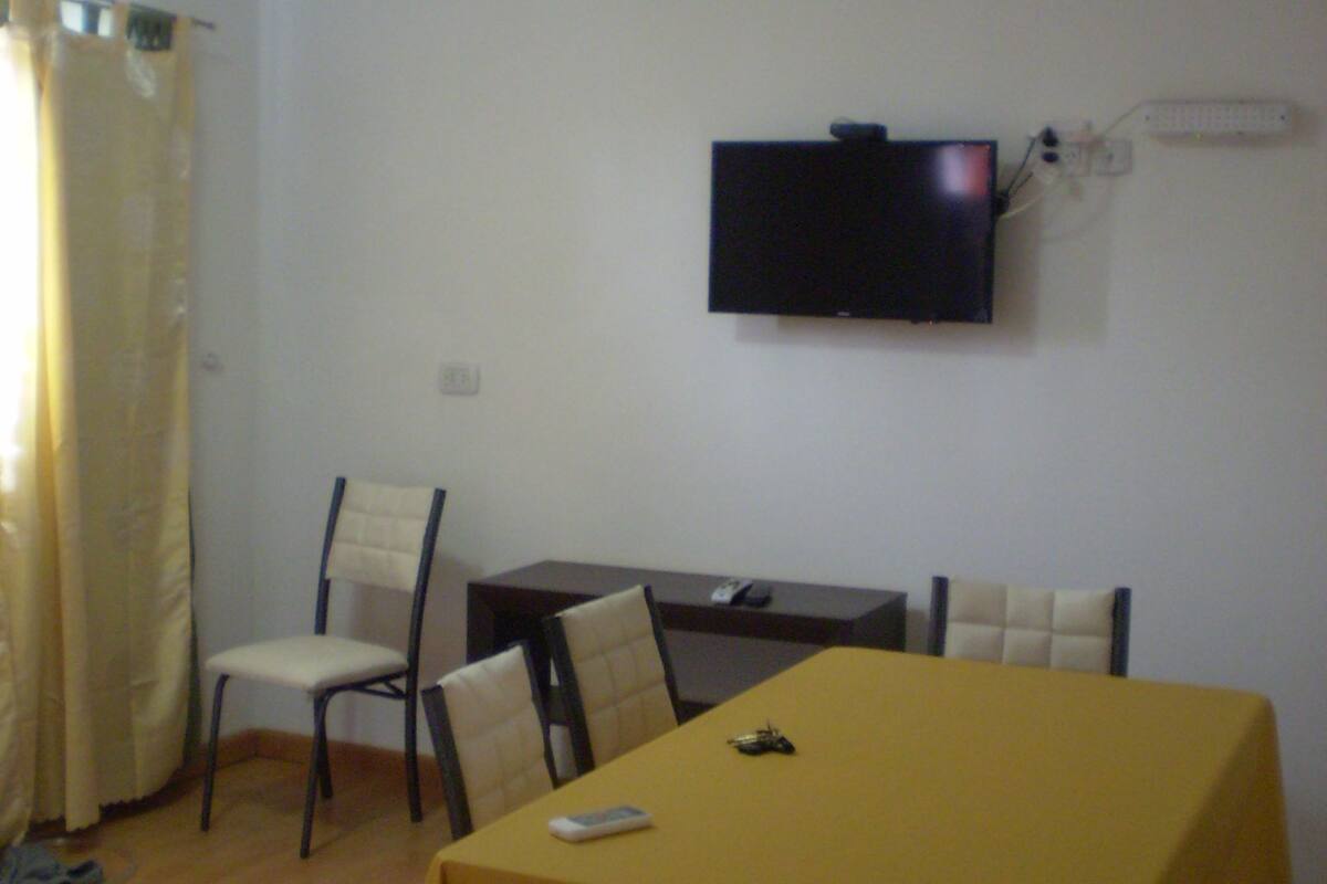Comedor con una mesa amarilla, cuatro sillas, un televisor de pantalla plana en la pared y un ventanal con cortinas.