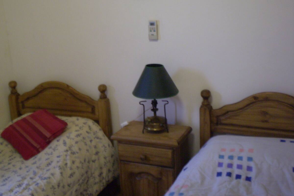 Un dormitorio con dos camas de madera, una mesita de noche con lámpara y un termostato fijado a la pared.