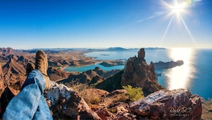 Uma pessoa sentada em um afloramento rochoso, com vista para uma paisagem costeira com montanhas e um corpo d'água.